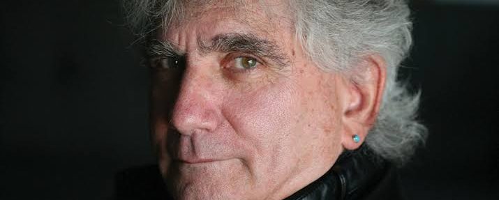 Joel looks at the camera from a side profile. He is wearing a dark shirt against a dark background. He has light skin and grey hair.